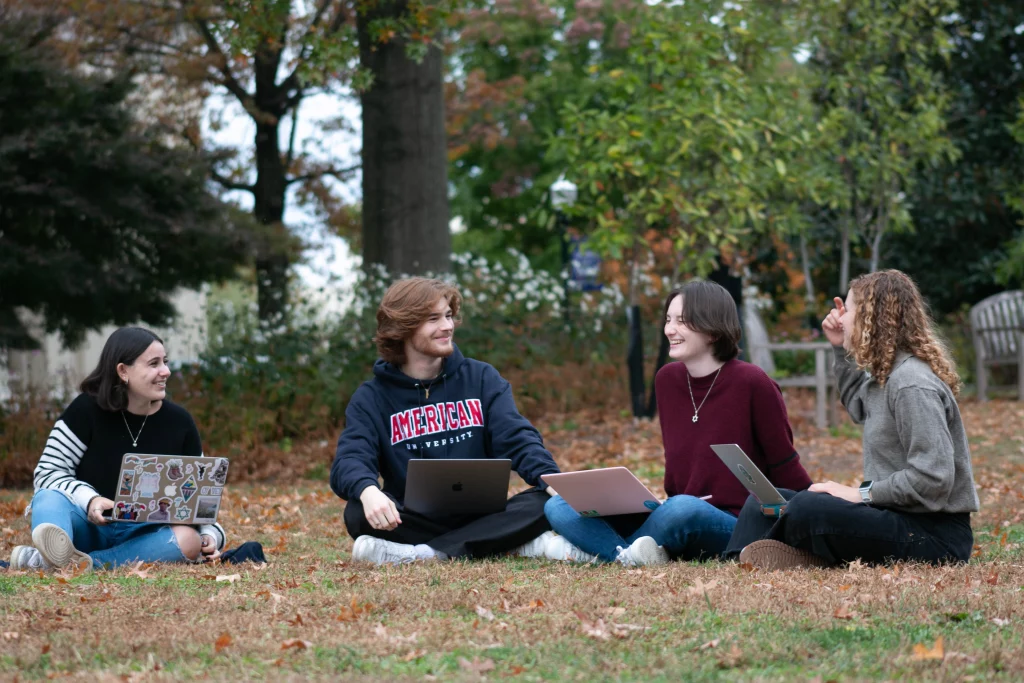 Students sit on a campus quad with their laptops, talking and laughing.