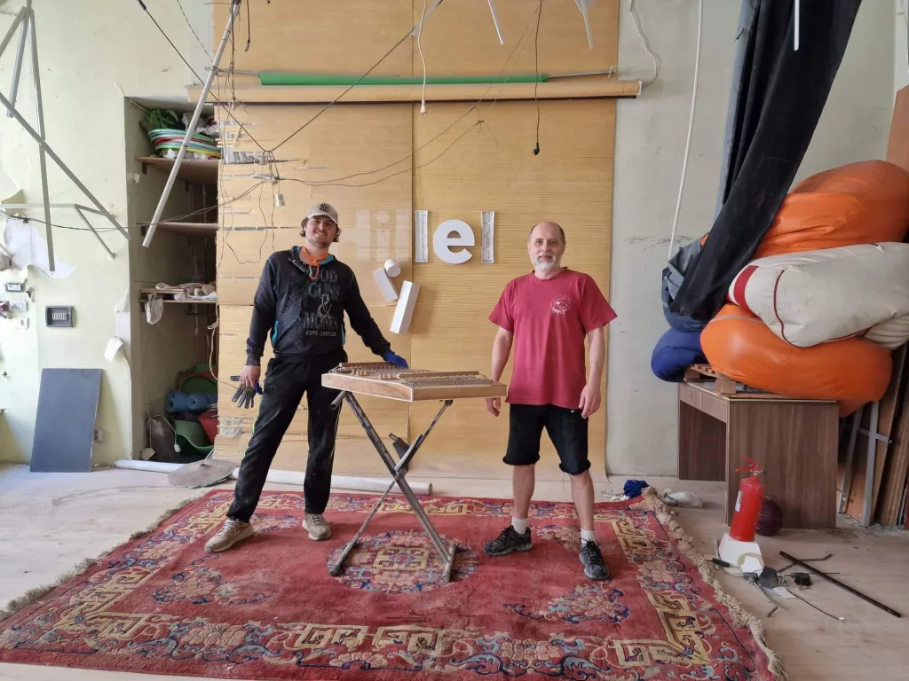 Two man stand at a workbench in front of a partially destroyed Hillel sign