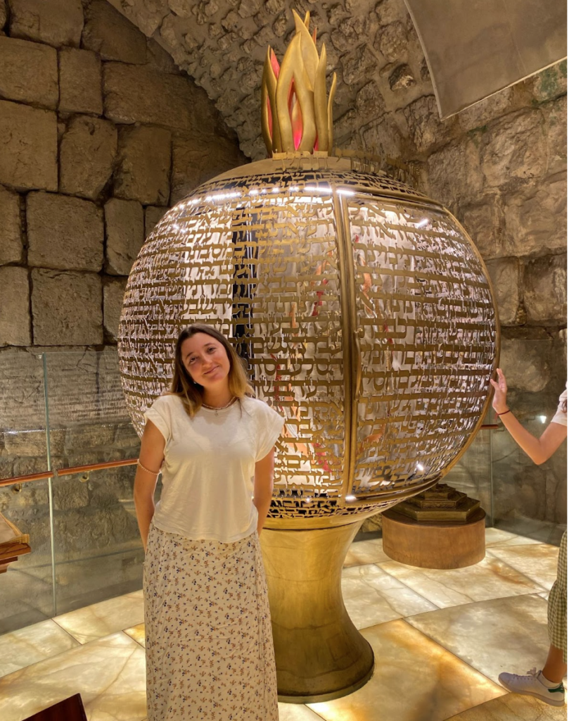 Student stands in front of a large gold sphere with Hebrew text inscribed on it