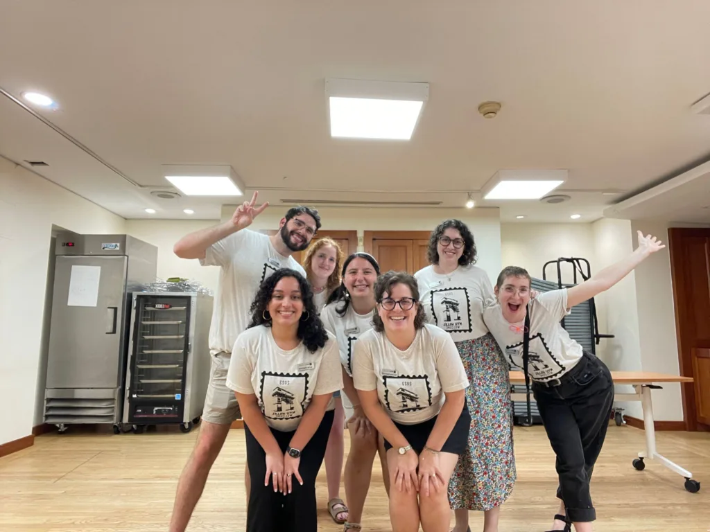 Welcome to NYU Hillel! Seven Hillel staff members wearing white and black shirts pose in an empty kitchen with big smiles and outstretched hands. 
