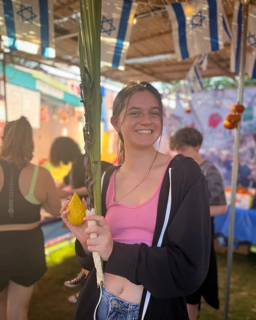 A student holds a lulav and etrog in a sukkah.
