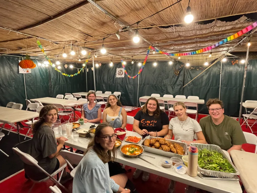 Students sit in a sukkah at a table covered with food.
